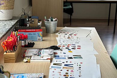 Auf einem Tisch liegen ausgedruckte Vorlagen mit Kleidungsstücken und Symbolen sowie verschiedene Bastel- und Schreibmaterialien wie Scheren, Stifte und Marker. Im Hintergrund ist ein heller Gruppenraum zu sehen.