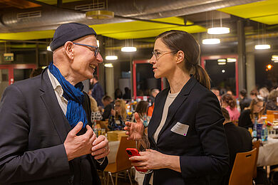 Oberkirchenrat Dietrich Bauer (links) spricht mit Vicky Felthaus (rechts). Er trägt einen dunklen Anzug, einen blauen Schal und eine blaue Mütze. Sie trägt einen dunkelblauen Blazer. Beide unterhalten sich angeregt.