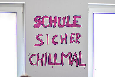 Auf einer Wand zwischen zwei Fenstern stehen in pinker, handgemalter Schrift die Worte „Schule Sicher Chill Mal“