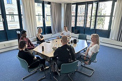 Mitarbeitende der Jugendberufshilfe sitzen gemeinsam mit Heike Praetz, stv. Geschäftsführerin des Jobcenters Leipzig, an einem Tisch. Alle Personen scheinen in ein anregendes Gespräch vertieft zu sein. Der Teppich ist blau, die Fenster im Hintergrund sind teilweise geöffnet.