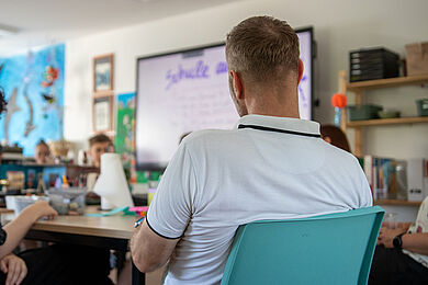 Eine Person wird von hinten gezeigt, wie sie an einem Tisch sitzt. Im Hintergrund sind unscharf der Tisch mit einigen Bastelmaterialien und mehreren weiteren Personen zu sehen. Dahinter sind eine Wandmalerei mit Fischen im blauen Wasser und ein Whiteboard mit dem Wort „Schule“ zu sehen.