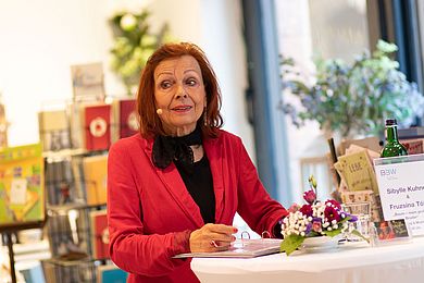 Sibylle Kuhne in einer Portraitaufnahme während sie liest. Sie steht an einem hohen Tisch, auf dem ein Hefter, Blumen, Wasser und ein Namensschild stehen.