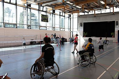 In einer Turnhalle sind Netze aufgespannt. Es wird Federball gespielt. Unter den Sportler*innen sind einige Personen im Rollstuhl und auch kleinwüchsige Menschen.