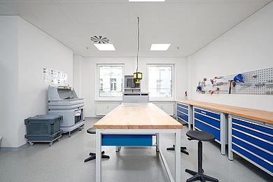 Blick in den Werkstattraum, an der rechten Seite befinden sich Werkbänke und Metallschubladen, an der Wand hängen Werkzeuge, in der Mitte befindet sich ein großer Tisch