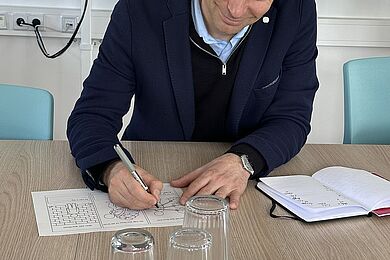 Eine Person sitzt an einem Tisch und bearbeitet Arbeitsblätter mit einem Stift. Vor ihr stehen mehrere umgedrehte Gläser, unter einem Glas befindet sich eine kleine grüne Figur.