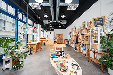 Raumaufnahme der Elsterkammer, an der rechten Seite sind Regale mit Ware, in der Mitte steht ein niedriger Holztisch mit Artikeln, auf der linken Seite die Fensterfront