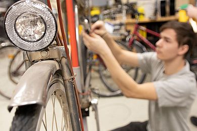 Ein Fahrrad ist im Vordergrund, im Hintergrund repariert ein Jugendlicher das Fahrrad