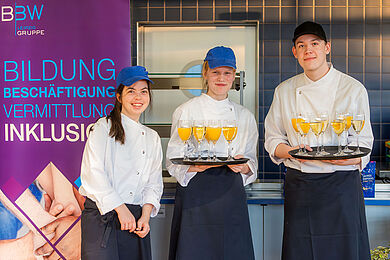 Neben dem Rollup der BBW-Leipzig-Gruppe stehen drei Auszubildende des Fachbereichs Ernährung des BBW Leipzig. Zwei von ihnen tragen ein Tablett mit Sekt- und Orangensaftgläsern. Alle tragen dunkle Küchenschürzen und weiße Kochblusen. Sie lächeln in die Kamera.