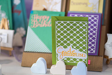 Grußkarten in verschiedenen Designs und Farben zu unterschiedlichen Anlässen. Im Vordergrund eine grün-weiße Karte mit der Aufschrift "Gute Besserung"