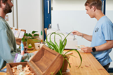 Zwei Personen stehen an der Kasse der Elsterkammer, im Vordergrund stehen eine Pflanze und eine Truhe