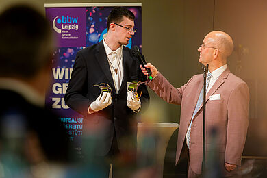 im Fokus des Bildes stehen zwei Männer. der linke Mann hat weiße Handschuhe an und hält zwei Brückenschlagpreise in den Händen. Er wird von einem weiteren Mann, dem Moderator, interviewt. Der Moderator trägt einen altrosa Anzug.